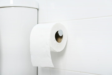 A white roll of soft toilet paper neatly hanging on chrome holder on a white bathroom wall. Close up