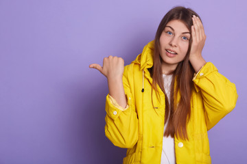 Closeup portrait of shocked woman dresses casual yellow jacket pointing thumb aside, keeping hand on head, lady with long beautiful hair against lilac wall. Copy space for advertisement or promotion.