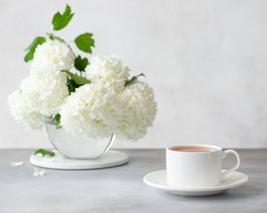 white cup of hot tea and a vase with a beautiful bouquet of white flowers. tea time. monochrome. horizontal image. light gray background with place for text
