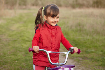 Obraz premium Outside picture of cheerful joyful little girl with pony tails, riding bicycle alone, having fun, breathing fresh air, being fond of nature, playing game, having delighted facial expression.