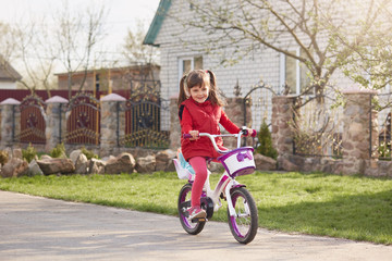 Adorable of cute female child riding bike on road near her house, charming girl wearing red clothing, happy kid walking in open air, smiling girl with her doll on bike. Childhood, children concept.
