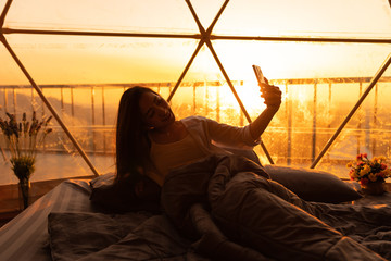 Cheerful woman is use smartphone on bed in tent around mountains under sunset light sky enjoying the pleasure and freedom Traveling along the mountains, freedom and active lifestyle concept.