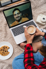 A young woman making a video call with her coworker at home while having coffee and eating a homemade cookie