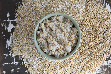 Top view of cooked barley porridge in green bowl on the wooden background with raw pearl barley and barley groats