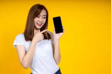 Naklejka premium Young elegant Asian woman smiling and pointing to smartphone isolated on yellow background