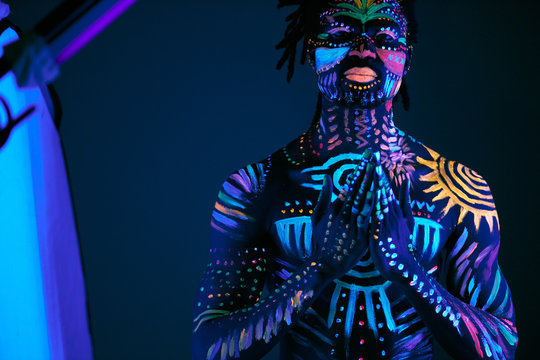 african prayer with UV body art glowing in darkness, colorful ethnic prints on naked skin. abstract fantastic futuristic paints, fluorescent make-up. man pray for the best