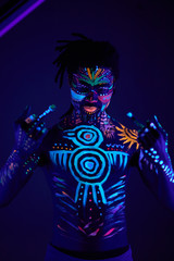 cool african boy with fluorescent make-up showing fuck gesture at camera, fearless man isolated in dark studio, luminescent paint glows in neon lights
