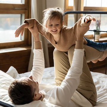 Square photo happy father playing with little daughter in bedroom, lying in bed, loving caring dad carrying preschool girl pretending flying with hands outstretched, family playing funny game