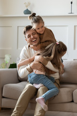 Funny little girls hugging cuddling smiling laughing father sitting on comfortable couch in living room, excited father having fun with two adorable little daughters, enjoying free time together - obrazy, fototapety, plakaty