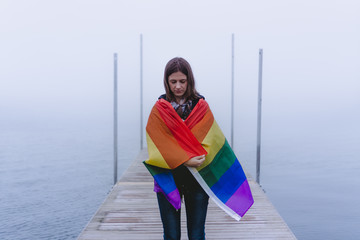 Beautiful woman standing on a pier wrapped in an LGBT flag.Woman who supports the rights of minorities, gays and lesbians. The woman shows solidarity with the LGBT community. Beautiful moody frame.