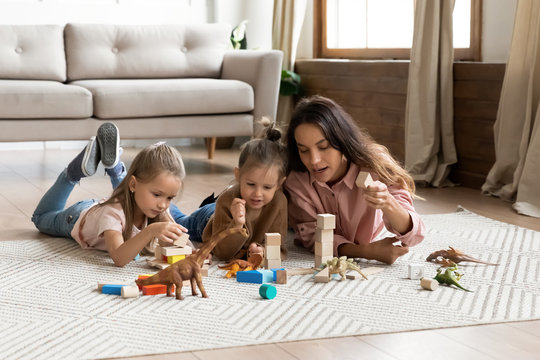 Young mother playing with two little daughters, lying on warm floor in living room, loving mum spending weekend with children at home, cute girls with baby sitter playing with wooden blocks