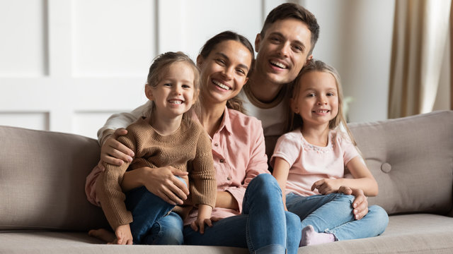 Portrait of smiling mother and father with two little daughters sitting on couch, looking at camera, happy parents hugging two preschool girls sisters, large family posing for photo at home