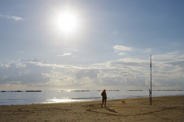Woman walking with a dog on the beach, the sun shines high.