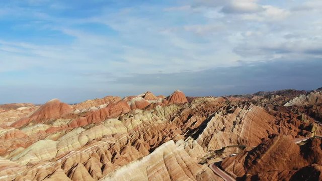 Aerial Top View of Rainbow Mountains Geological Park. Stripy Zhangye Danxia Landform Geological Park in Gansu Province, China. Flying Drone Picture of Tourist Buses on a Road in Valley on a Sunny Day.