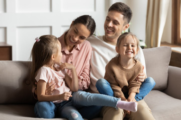Happy young mother and father with two little daughters sitting on couch, looking at each other,...