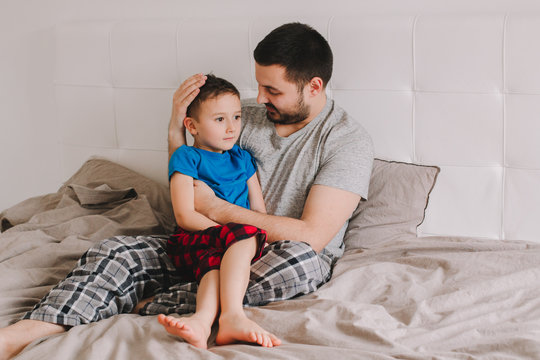 Caucasian Father Talking To Boy Son. Man Parent Hugs Kid Child On Bed In A Bedroom At Home. Authentic Lifestyle Real Family Candid Moment. Happy Fathers Day Holiday. Serious Talk.