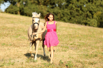 Young girl in pink dress run with pony through meadow in sunny summer afternoon