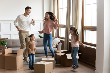 Fototapeta na wymiar Happy family with two little daughters celebrating moving day, excited by relocation into new house, laughing mother and father with children jumping in living room with cardboard boxes