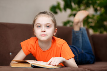 Cute caucasian little girl reading book while lying on sofa at home during quarantine due to coronavirus. Education and leisure concept
