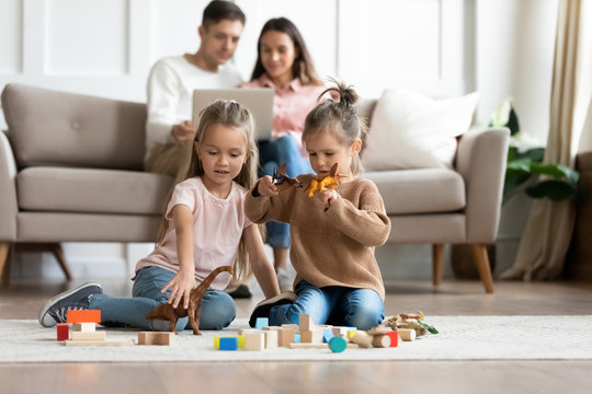 Two little girls sisters playing with toys while parents resting on couch in living room, cute small daughters sitting on warm floor at home, having fun together, friendship between siblings