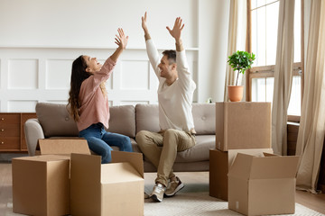 Happy young couple raising hands, sitting on couch in living room with cardboard boxes, celebrating...