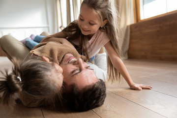 Happy father lying on warm floor with two adorable little daughters close up, hugging and cuddling, family having fun at home, spending free time together, cute preschool girls playing with dad