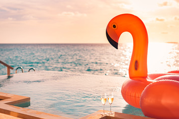 Luxury infinity swimming pool. Sunset holiday travel vacation background with funny flamingo toy float header an champagne glasses as romantic couple background, exotic love romance destination.