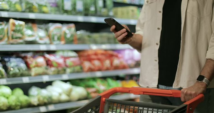 Crop view of guy holding phone, typing and touching screen in supermarket. Man using smartphone while walking and pushing shopping cart near green grocery shelves. Blurred background