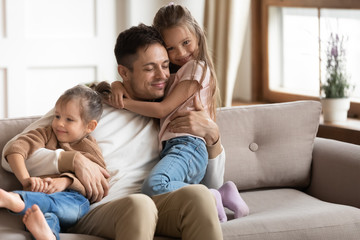 Loving young father cuddling with two little daughters, sitting on cozy couch at home, family...