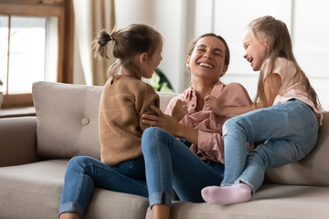 Happy mother playing with two little daughters, sitting on couch at home, cute preschool girls tickling laughing mum, family having fun, spending weekend together, playing funny game