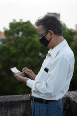 Fototapeta premium Senior man with corona preventive mask operating calculator standing on rooftop outdoor. Covid, Lock down and Home isolation