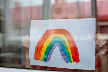 Little girl in the window holds a picture of a rainbow symbolizing the imminent end of the quarantine in connection with the coronavirus pandemic.