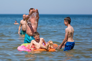 Zabawa rodziny w morzy, ojciec polewa wodą chłopca na kolorowym dmuchanym materacu - obrazy, fototapety, plakaty