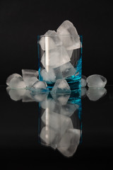 Still-life of a blue glass filled with ice cubes, the composition is mirrored on a dark glossy top.