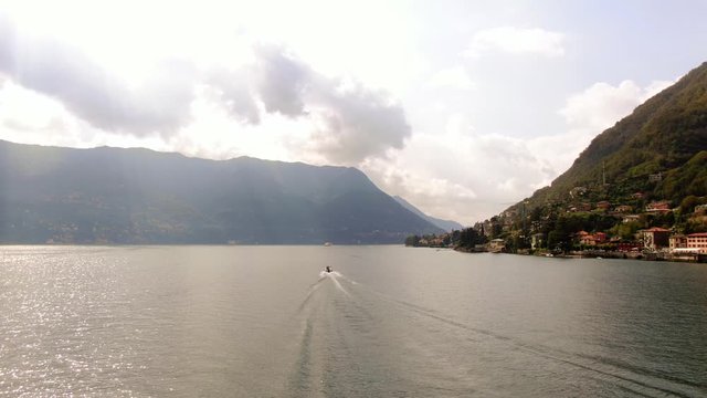 A motor boat floats past the village of Laglio towards rays of sun, which break through clouds against the backdrop of the Italian Alps. Aerial view. Shooting from a drone on a summer day on Lake Como