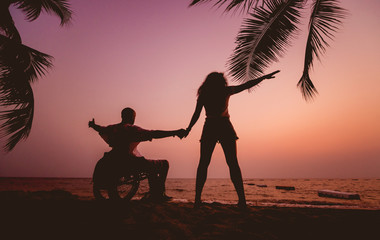 Fototapeta na wymiar Disabled man in a wheelchair with his wife on the beach. Silhouettes at sunset