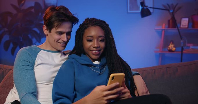 Millennial couple smiling and looking at phone while spending time. Handsome afro american girl scrolling smartphone while sitting in her boyfriend embrace on sofa at home