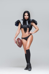 Full length portrait of sexy brunette female american football player in uniform posing with a ball isolated on grey background