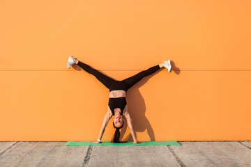 Overjoyed happy girl with perfect athletic body in tight sportswear doing yoga handstand pose with spread legs against wall and showing tongue, having fun. Gymnastics for body balance, flexibility