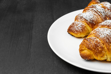 Delicious buttery croissants on a white plate on a black wooden background with copy space.