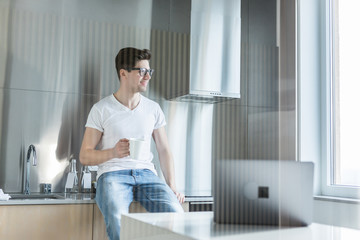 Fototapeta na wymiar Young handsome man having coffee while talking on the phone in kitchen at home