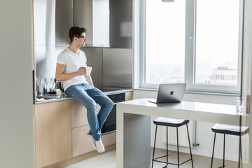 Young handsome man smiling and holding coffee at home in the kitchen
