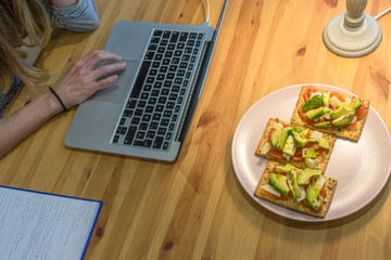 
young person working on a laptop while having toast with tomato and avocado on wooden table