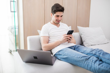Fototapeta premium Young handsome man sitting relaxed on sofa with laptop checking his smartphone messages