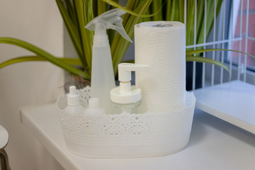 Basket with anti-bacterial protective equipment. Antiseptic bottles, antiviral jars, a clean roll of paper. Stands on the windowsill against a background of flowers.