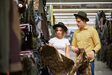 young man and woman in search of equipment for hunting in store, choose sportive wear with green swamp prints, stand in hats, share opinions