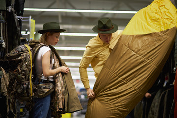 Fototapeta premium young good-looking married couple keen on travel, they choosing the best variant of tent in sportswear store, wearing hats and bags for travelling on their backs
