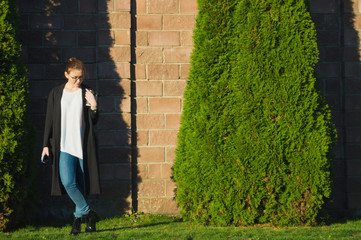 Woman standing in thoughtful pose  holding cup of coffee and smartphone. Full length outdoor...