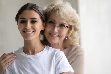 Close up headshot portrait picture of happy mature mother hugs from behind adult daughter enjoying tender moment looking at camera. Smiling woman and older mum embracing having fun together.