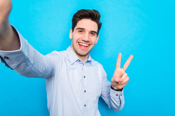 Handsome smiling young man is making selfie on smartphone and gesturing peace on blue background